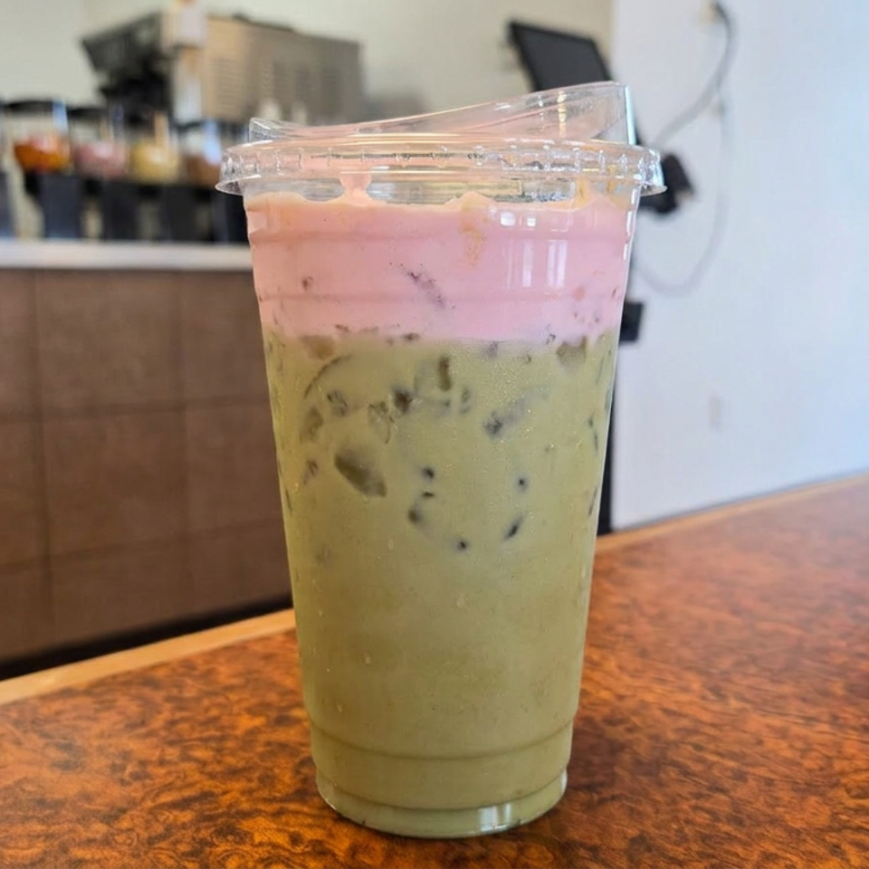Layered iced matcha drink topped with pink cream in a clear cup on a wooden café table.