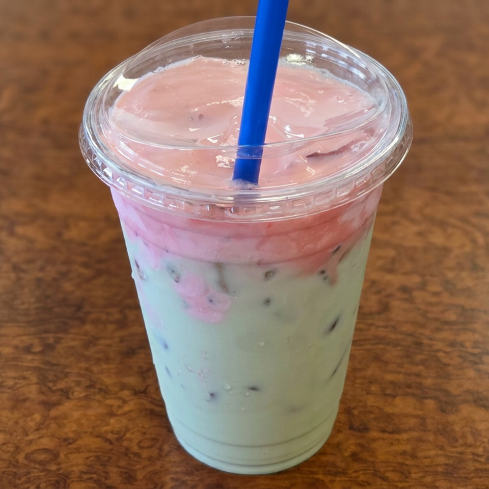 Pink and pale green iced beverage with a blue straw in a clear plastic cup on a wooden table.